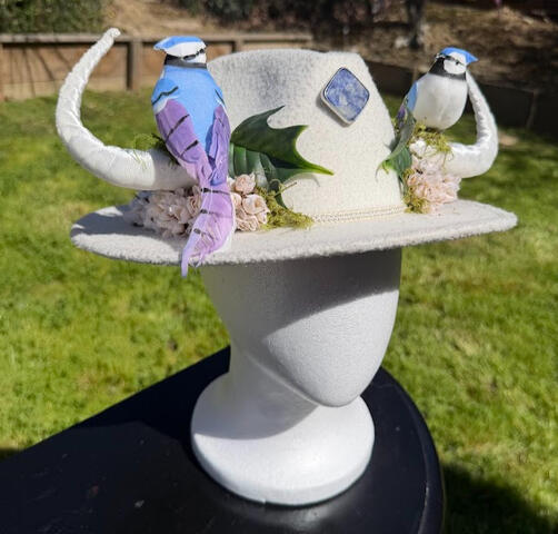 White wide brim hat with velvet cream white faun horns. A blue jay perches on each horn surrounded by white flower clusters, light green sprouts, and moss.