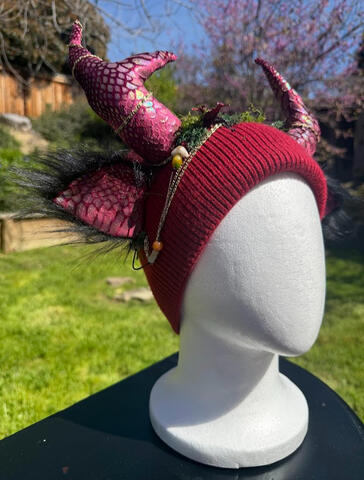 Dark red beanie with iridescent maroon scaley antlers. Ears have black furred backing. Brass chains dangle from the side and curl up the antlers. A strand of jade beads hangs from the base of the antlers to the ears.