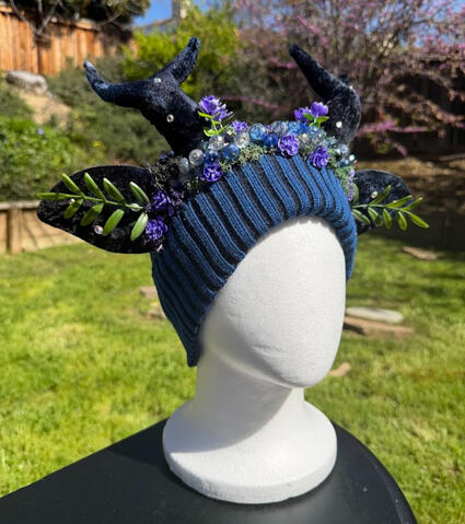 Deep blue beanie with small navy blue antlers with matching blue ears. A crown of iridescent beads line the forehead of the beanie, accented with moss and purple blossoms.