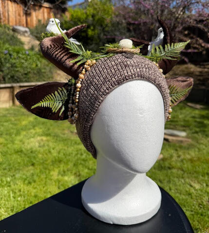 Mixed brown heathered beanie with brown velvet antlers. On each antler is a white bird surrounded by green moss. Green ferns sprout from either side, surrounding a small bird's nest at the center.