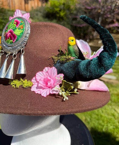 A golden budgie and gold-grey-green budgie perch on each horn. A beautiful round embroidered floral piece accents the front of the hat with dangling silver bells for a light jingle. Horns are poseable.