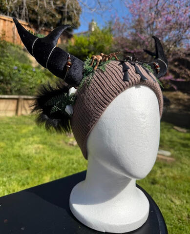 Brown to dark brown ombre beanie with black velvet antlers. Matching black furred ears. Two tarantulas painted in natural hues of tan and brown perch at the top of the beanie.
