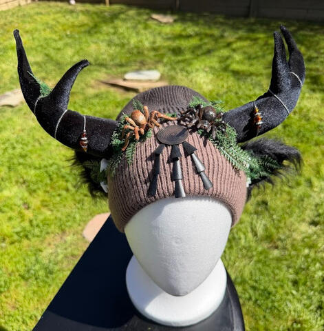 A mixed metal jewelry spiral piece adorns the forehead for a unique accent. Green moss and ferns sprout around the beanie with a few small white teeth for that spooky touch.