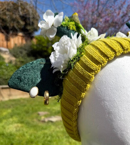 Lime green-yellow beanie with dark green velvet antlers. A green & yellow budgie perches on each antler, surrounded by moss and white flowers. On each ear are two earrings of gold and white for an elegant accent.