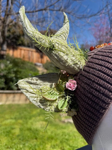 Light &amp; dark brown heather beanie with pale sage green antlers. Gentle light green ferns wrap around each antler with small light pink rose accents. A deep amber jade pendant &amp; beaded chain adorn the front of the beanie for a natural flare.