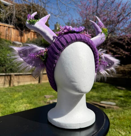 A light purple flower blooms from the center, surrounded by flowers and moss. A silky blue and purple butterfly perches on the flower. Horns are poseable, ears are pierceable & bendable. Beanie is lined inside with plush material for xtra comfort.