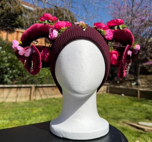 Maroon beanie with dark red faun horns. Hot pink roses adorn each horn with fluffy green moss. Matching dark red ears have a light pink silky butterfly and 2 unique accent earrings.