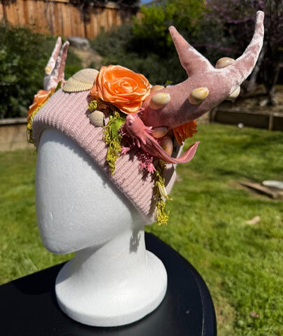 Light pink beanie with velvet pink antlers. Small seashells adorn the antlers and large shells decorate the base. A pink axolotl curls around either side of the beanie alongside bright salmon-pink roses.