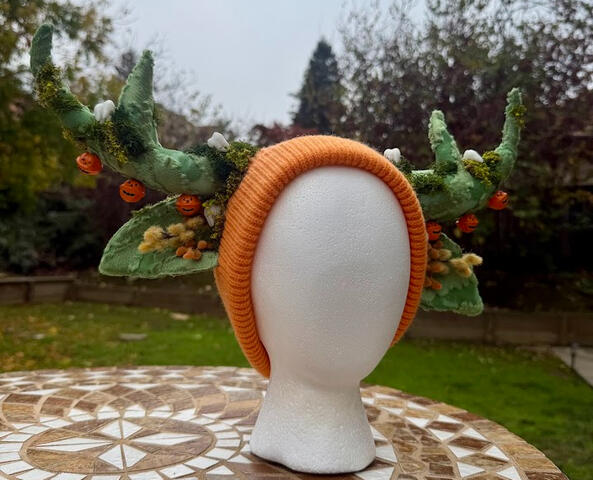 Mossy green antlers on an orange (unlined) beanie. Faux moss, white teeth, and autumnal plants decorate the antlers. 3 jingling orange jack o&#39; lanterns line each antler. Check out the video to hear them chime!