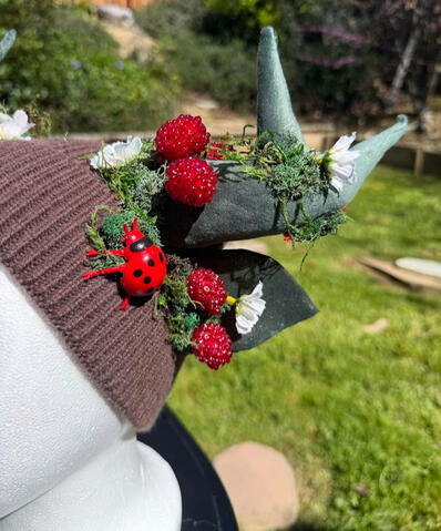 Brown beanie with green antlers and matching green ears. Raspberries and white flowers bloom from the base of the antler and ears. A round ladybug hugs each side of the beanie. Perfect for frolicking in the fields and meadows!