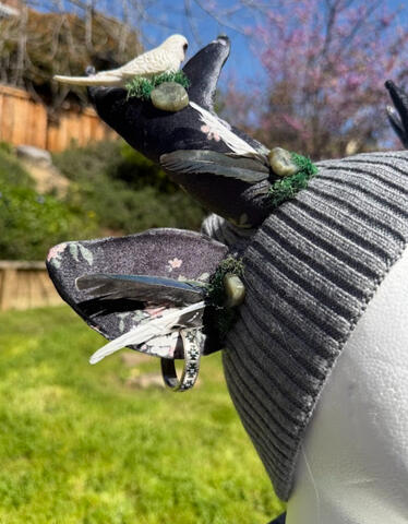 Grey beanie with floral patterned dark grey antlers. A white budgie and white/grey/blue budgie perch on each antler, surrounded by natural pieces of labradorite stone and moss.
