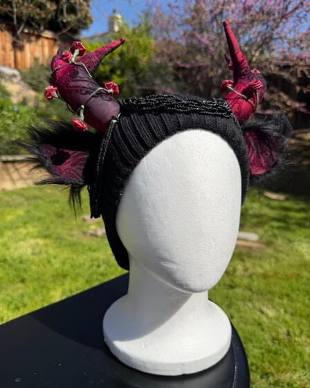Black beanie with mesh red & black velvet patterned antlers with matching ears. Strands of mini roses curl around each antler (they are poseable and removeable!) while strands of black beads cascade around the ears.