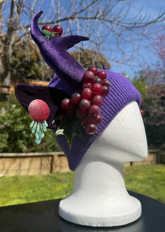 Purple beanie with velvet purple antlers. Red grapes drip from either side of the beanie, juicy enough to pluck and eat. Purple velvet ears are adorned with brass painted earring & acrylic accents. Horns are pose-able, ears are pierceable and bendable.