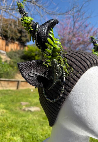 Dark grey beanie with textured scaly black antlers and matching ears. Bright green fern fronds curl around each antler along with a silver, black, and green magnetic beaded strand.