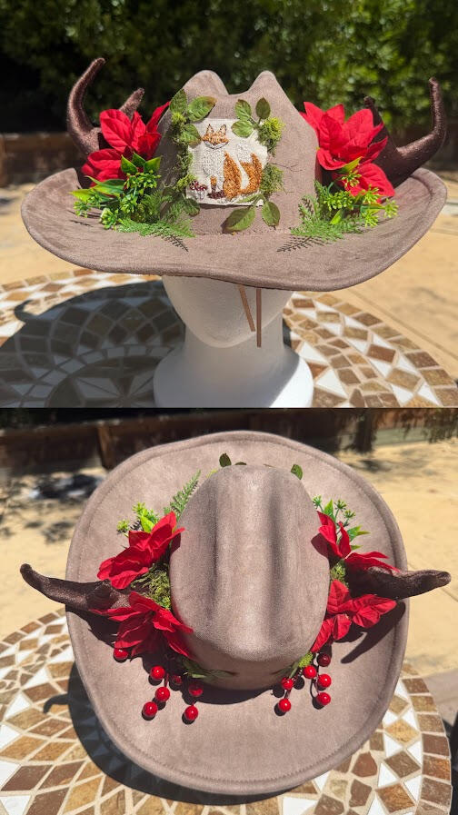 Upcycled grey-brown cowboy hat~ material assumed suede. Features brown poseable antlers with bright red flowers, berries, and green fern decor. An embroidered fox patch adorns the front of the hat.