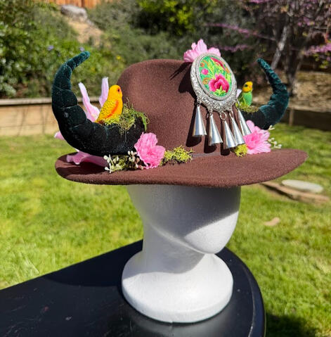 Chocolate brown wool hat with velvet green horns. Pink calla lilies & tiger pink lilies bloom out from the horns around the rim of the hat with light green-white sprouts.