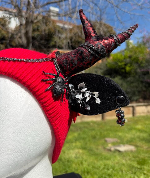 Bright red beanie with black & red metallic textured antlers. A black spider perches from either side near sprouting black-to-white ombre ferns. Slim furred black ears have rhinestone earrings that dangle with a shine.