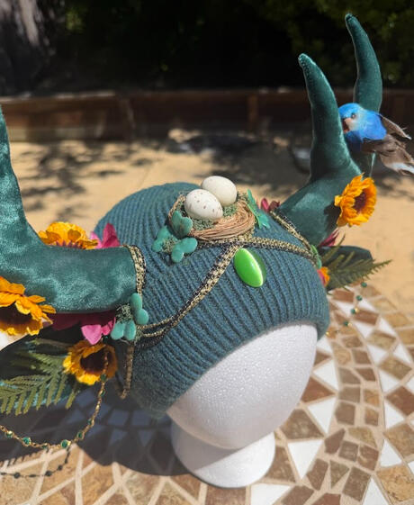 A bird's nest perches at the center of the beanie with a bluebird perched on either side on each antler. Green leaf clovers, sunflowers, fuschia flowers, and ferns adorn the beanie.