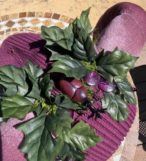 A big Japanese rhinoceros beetle sits at the top of the beanie surrounded by faux oak leaves and moss. Silver and purple beaded jewelry dangles from either side of the beanie.