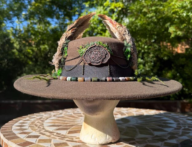Rim of the hat encompassed by mixed (semi-precious) stone cylindrical beads & black circle beads. A wooden pendant with burned floral design framed the center of the hat with brass bead strands & faux moss.